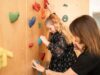 Mädchen klettert am Kletterwand im Behandlungsraum, Therapeutin unterstützt sie dabei