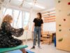 Kind schaukelt in Behandlungsraum, Therapeutin steht daneben